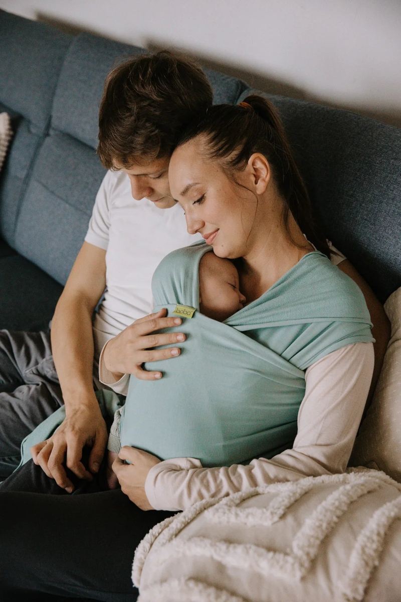 Una pareja en el sofá con su bebé recién nacido en un fular portabebés